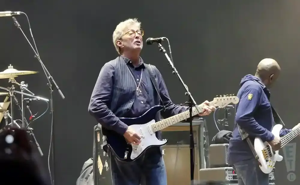 Guitarist Eric Clapton sings and plays a black Fender Stratocaster on stage with a bassist.