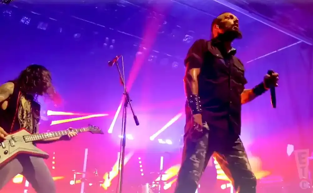 A male rock singer with a microphone stands center stage in front of purple and pink lights, while a guitarist plays to his left.