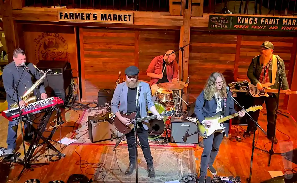 A band plays on stage, with a saxophonist/keyboardist, guitarist, drummer, and two bassists.