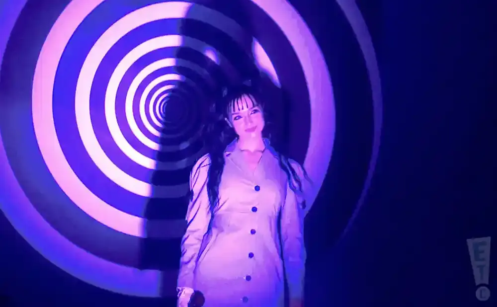 Woman in a light-colored button-down dress stands in front of a purple and white spiral background.