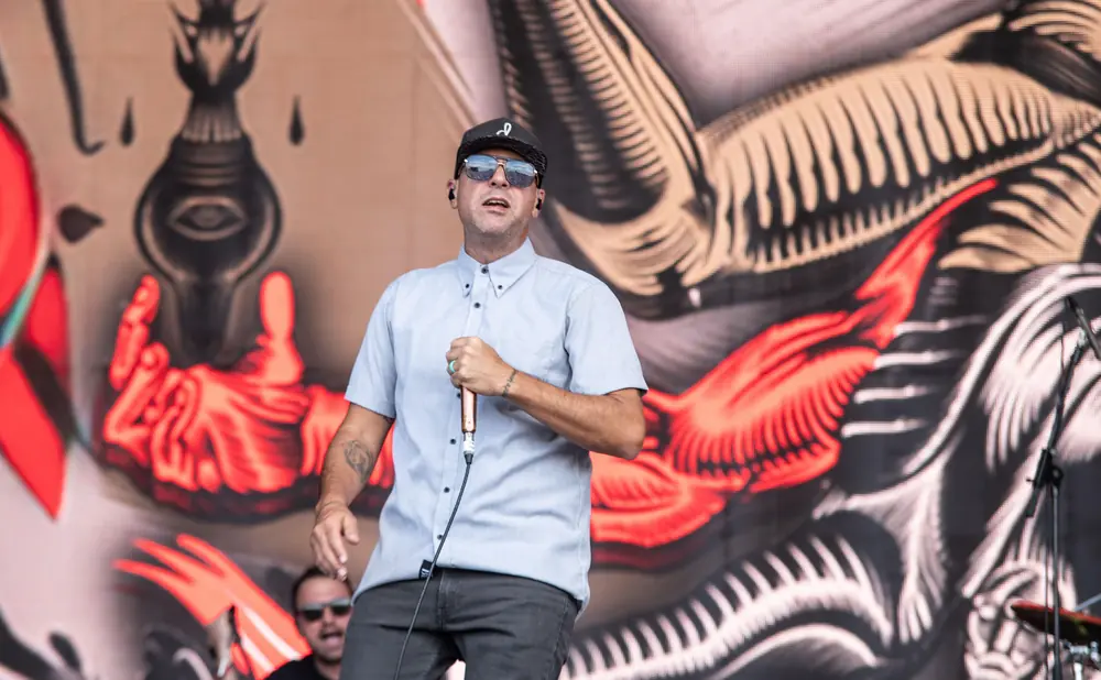 A man in a hat and sunglasses sings into a microphone on stage.