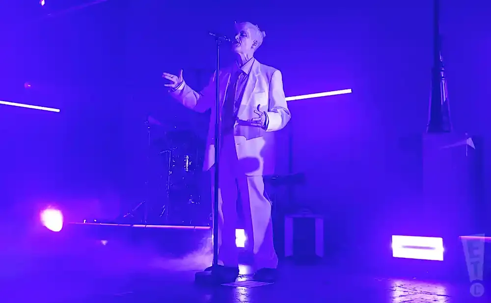 Singer in a white suit performing on a stage bathed in purple light.