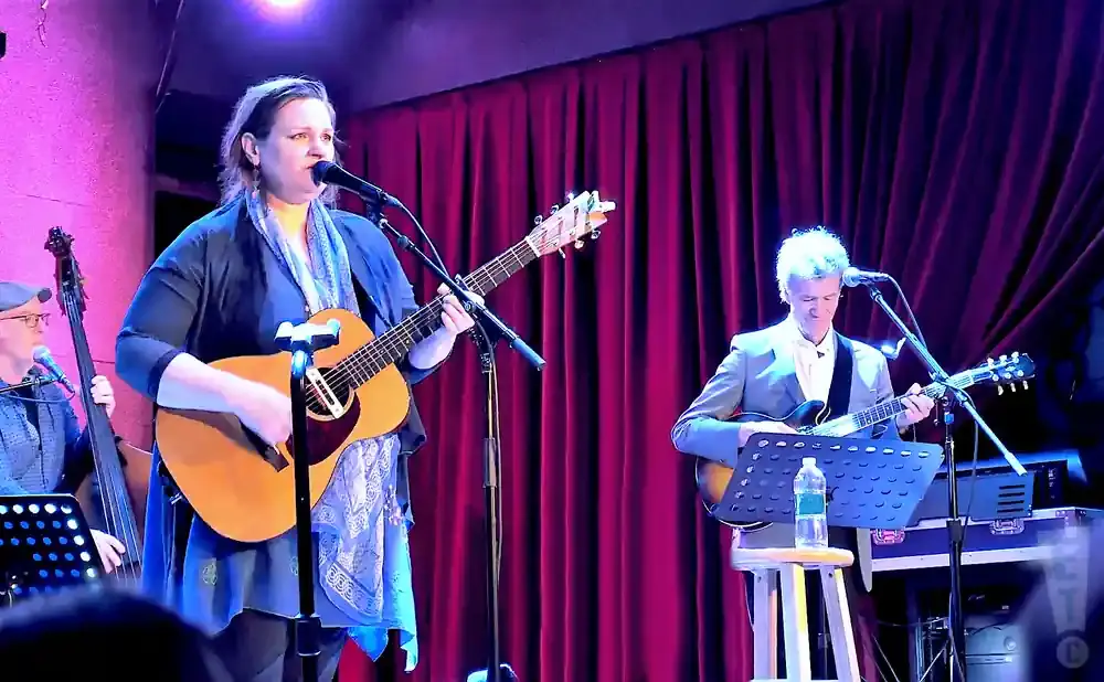 A woman plays guitar and sings into a microphone at a concert, with two men playing instruments behind her.