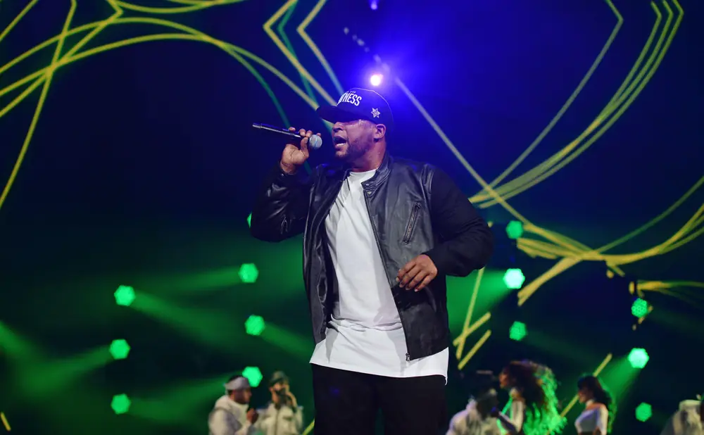 A male singer in a black leather jacket and white t-shirt performs on stage with a microphone.