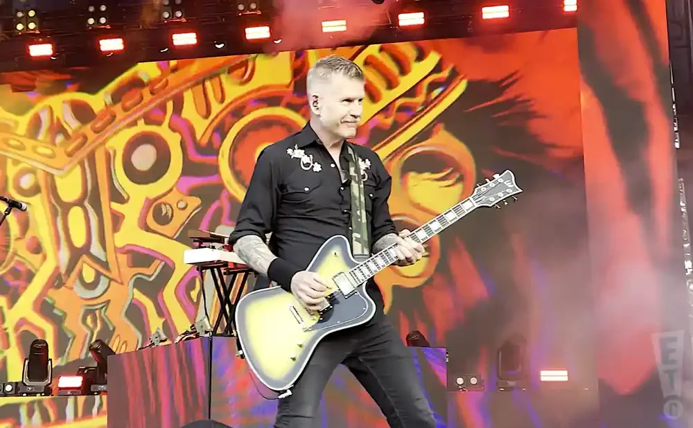 A man plays a yellow and grey electric guitar on stage with a vibrant, abstract background.