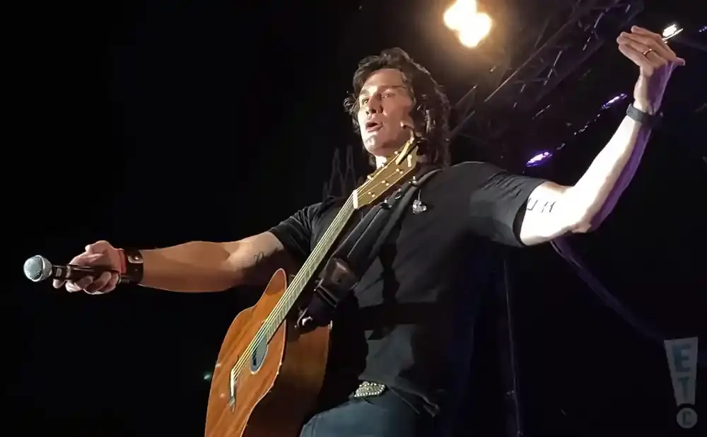 A male singer with long dark hair holds a microphone and plays an acoustic guitar on stage, wearing a black shirt and jeans.