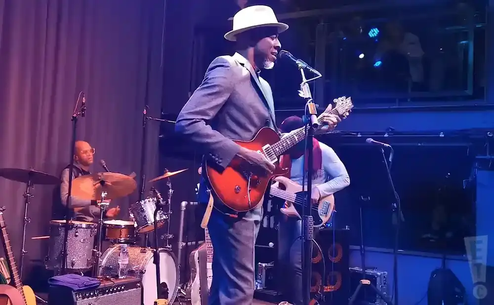 A man in a hat and suit plays an electric guitar on stage with a drummer and bassist.