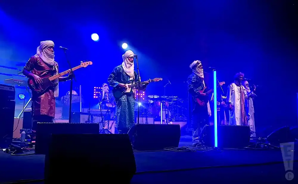 A band plays on a stage lit by blue lights. They wear traditional Tuareg attire and play guitars and bass.