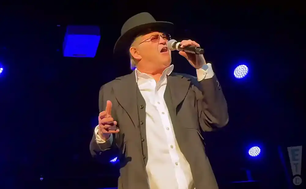 Singer in a black hat and suit, holding a microphone and gesturing with his hand, with blue stage lights in the background.
