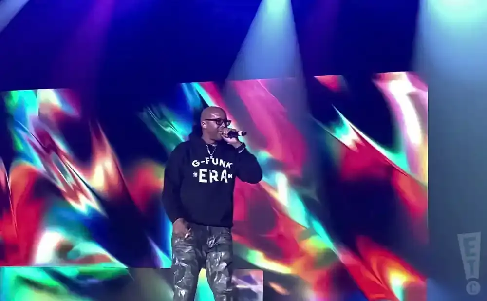 A man in a black hoodie and camouflage pants raps into a microphone on a stage with a colorful, abstract background.