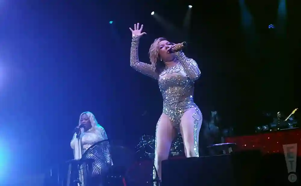 Two women perform on a brightly lit stage. The woman in front sings into a microphone, wearing a sparkly, form-fitting outfit.