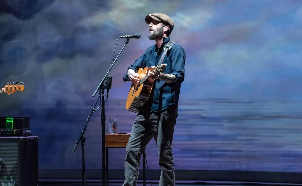 A man in a cap plays an acoustic guitar on stage with a blue, cloudy backdrop.