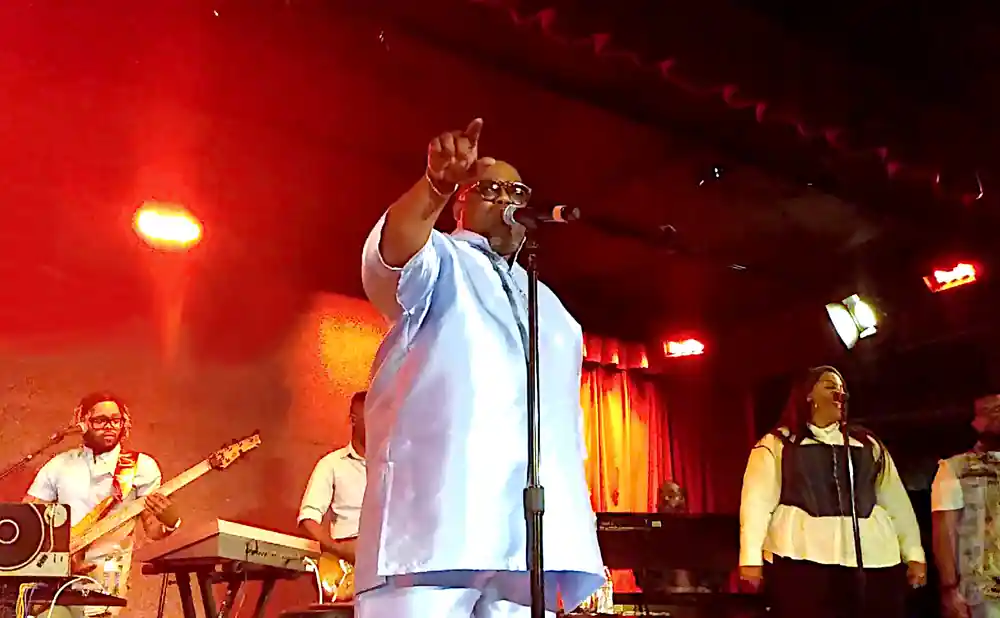 A male singer in a light blue outfit stands center stage with a microphone, pointing up at a concert.