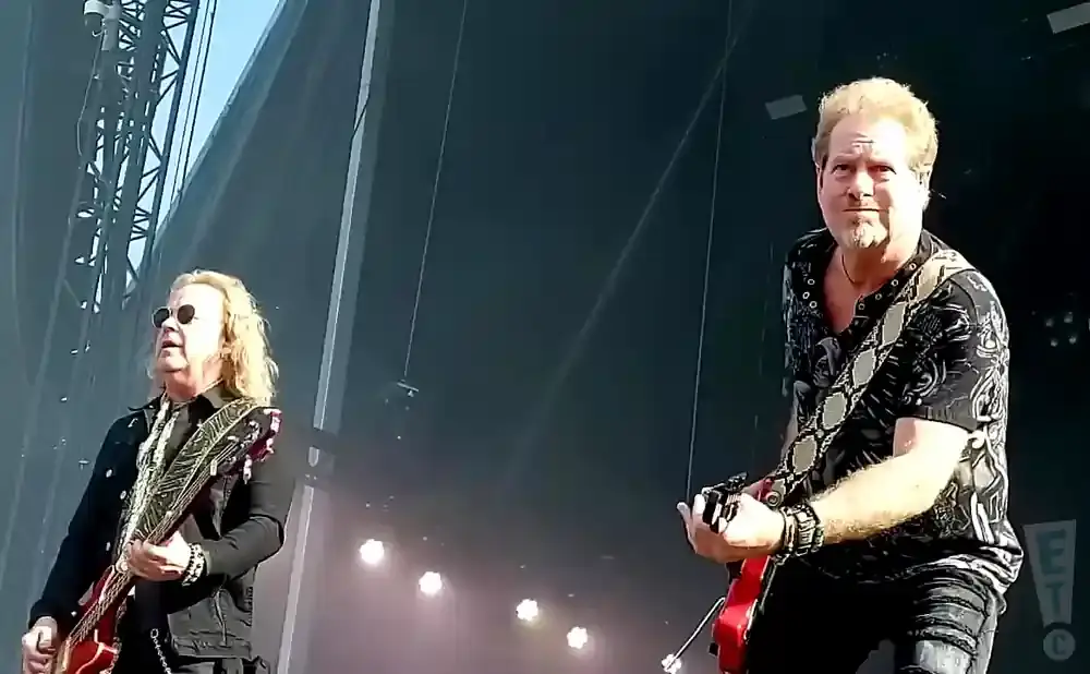 Two guitarists on stage, one with long blonde hair and sunglasses, the other with short blonde hair and a beard.
