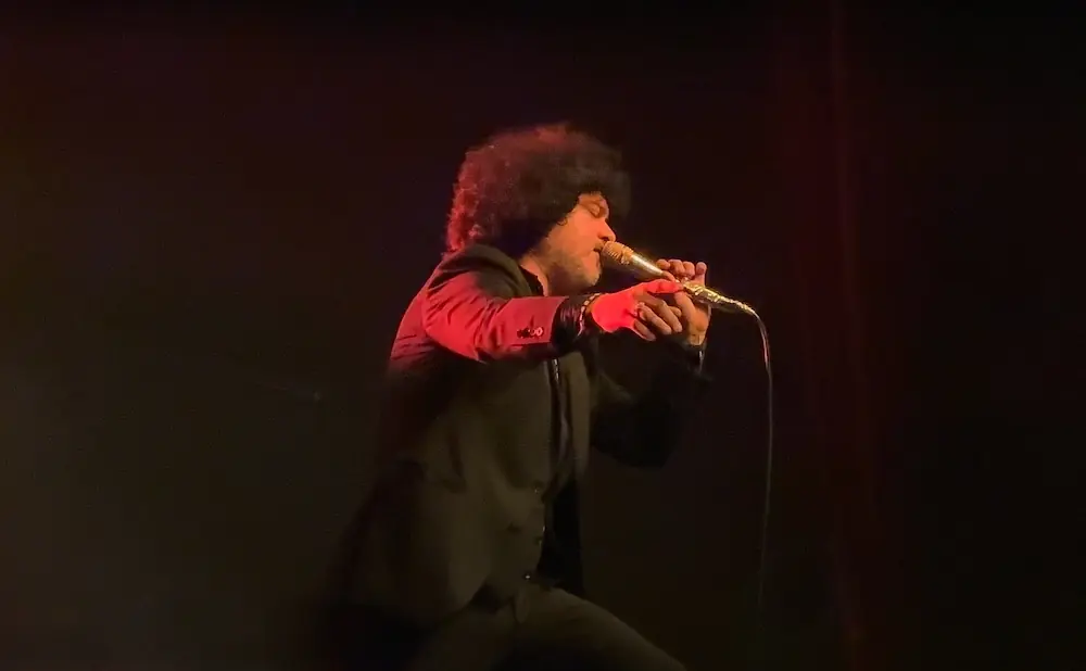 A man with an afro sings into a microphone on stage, wearing a black suit and red-lit jacket.