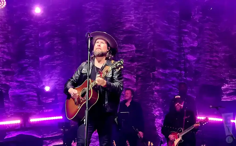 A man in a cowboy hat and leather jacket plays an acoustic guitar on stage with purple lighting.