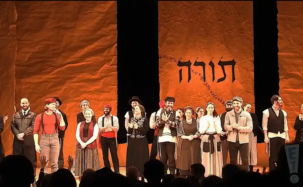 A group of actors in traditional clothing stand on stage in front of an orange backdrop with Hebrew text.