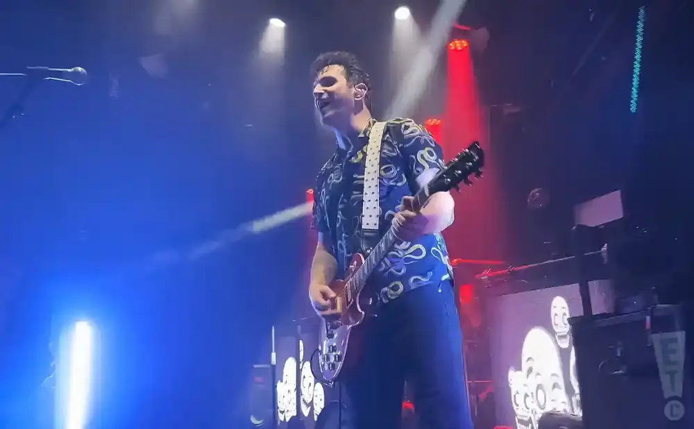 A male guitarist in a patterned shirt plays a red electric guitar on a brightly lit stage.