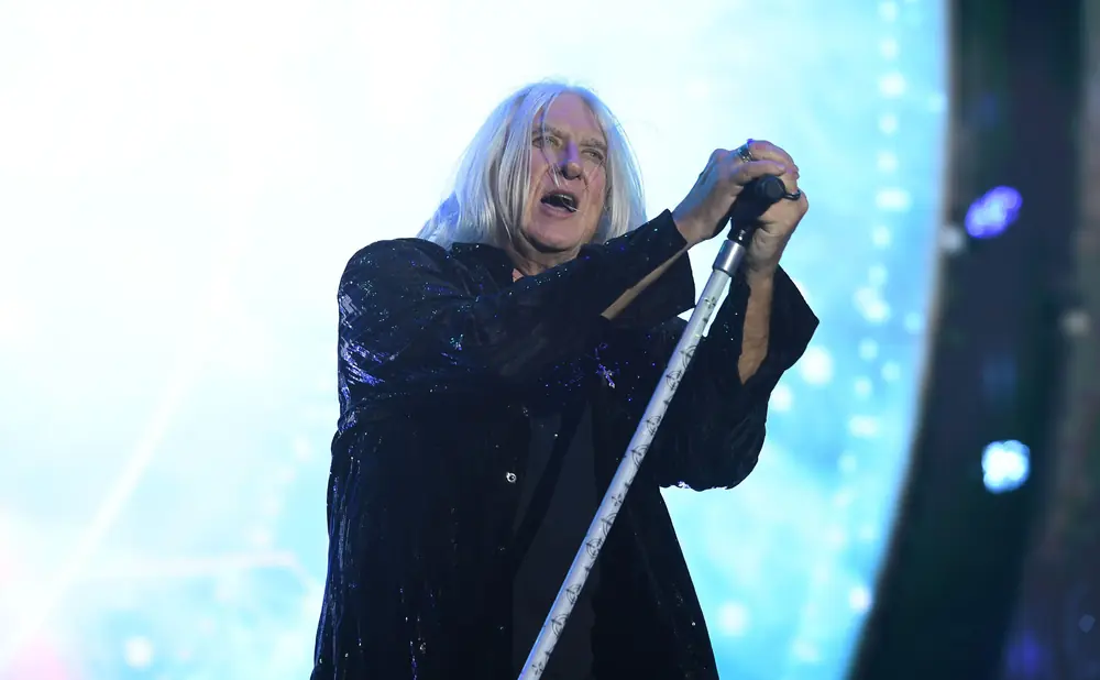 A man with long, white hair sings into a microphone, wearing a sequined jacket.