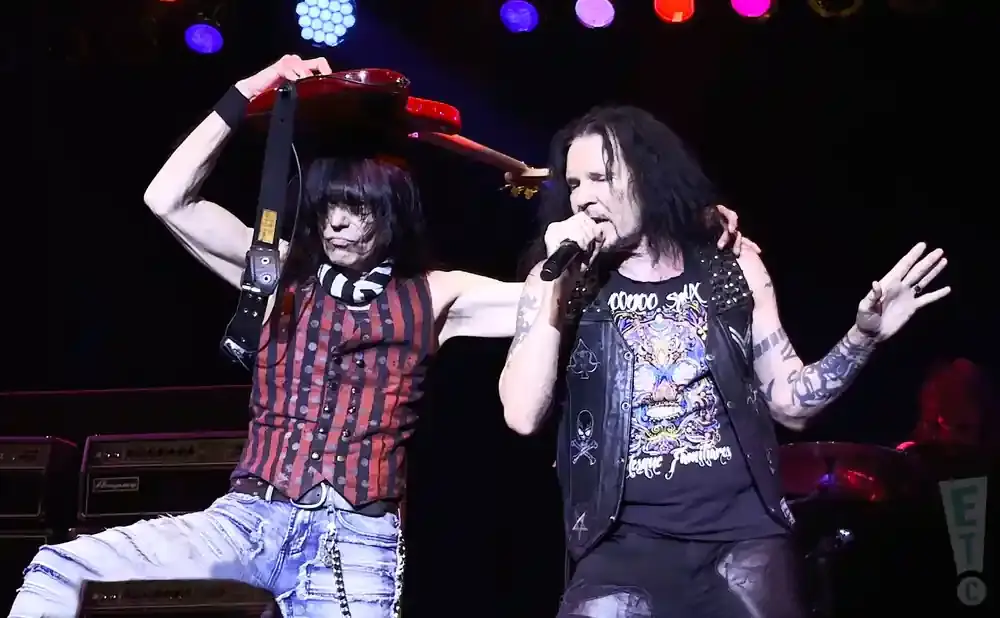 Two male rock musicians performing on stage, one holding a guitar above his head and the other singing into a microphone.