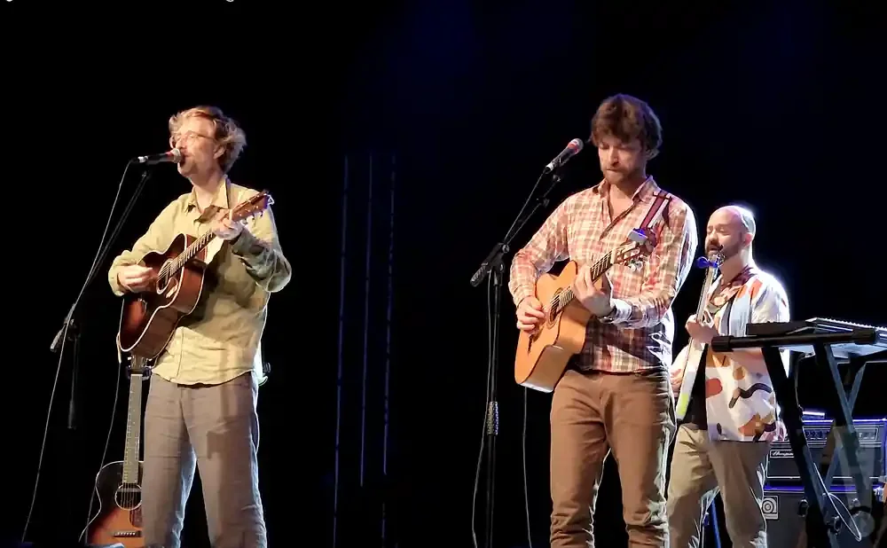 Three men play guitar and sing on stage. One plays an acoustic guitar and wears glasses.