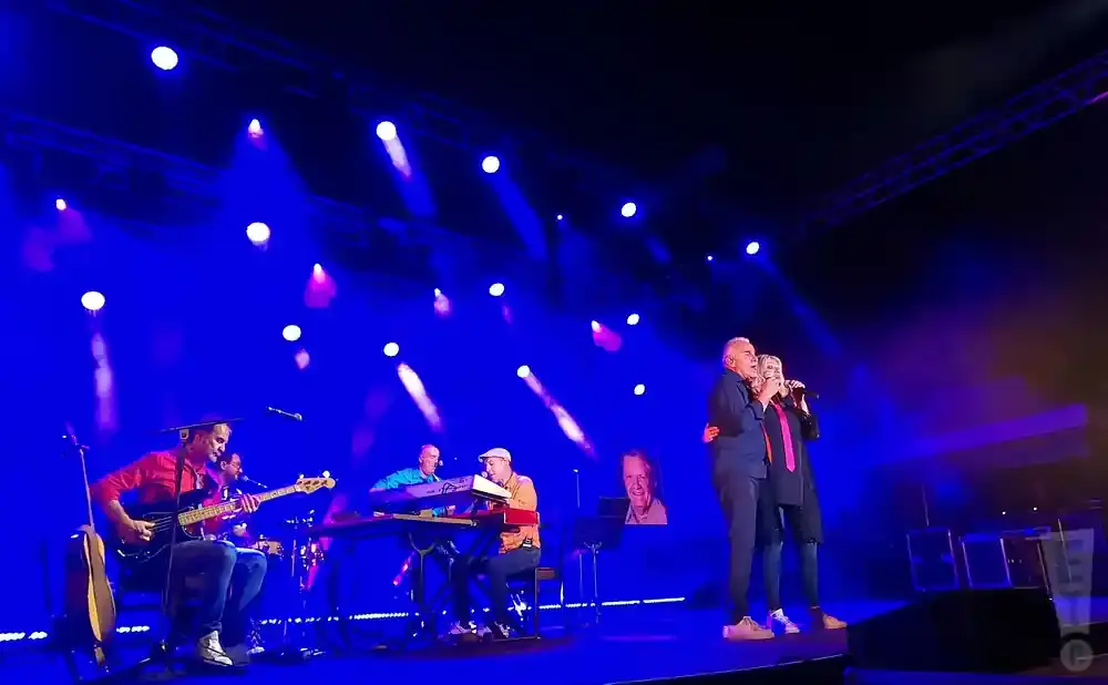 A band performs on a stage with blue lighting. Two singers stand in the foreground.