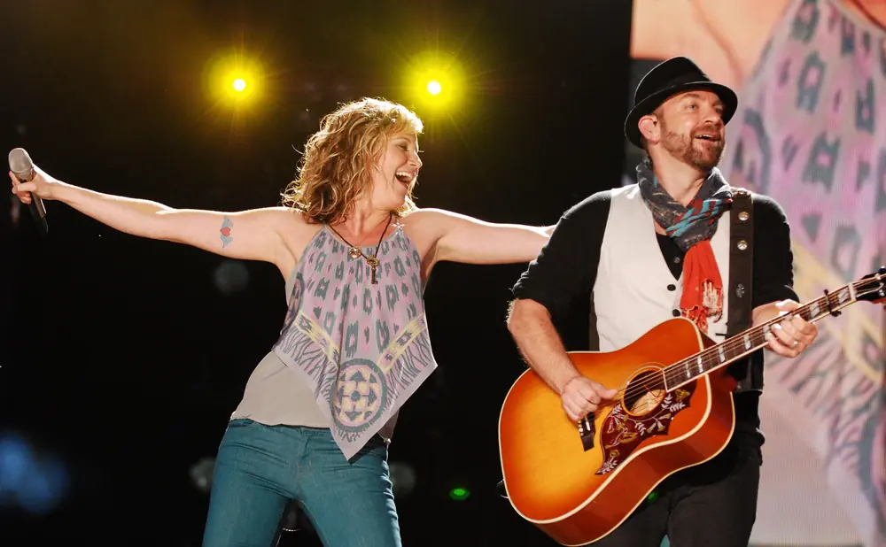 A woman sings into a microphone with her arms outstretched while a man plays an acoustic guitar onstage.