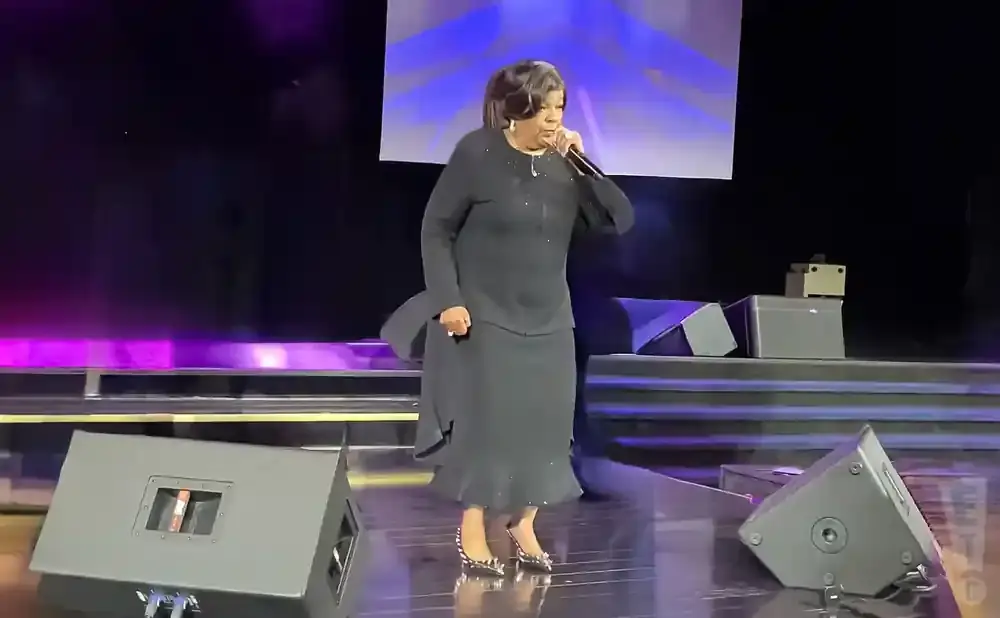 A woman in a black dress sings into a microphone on a stage with purple lighting.