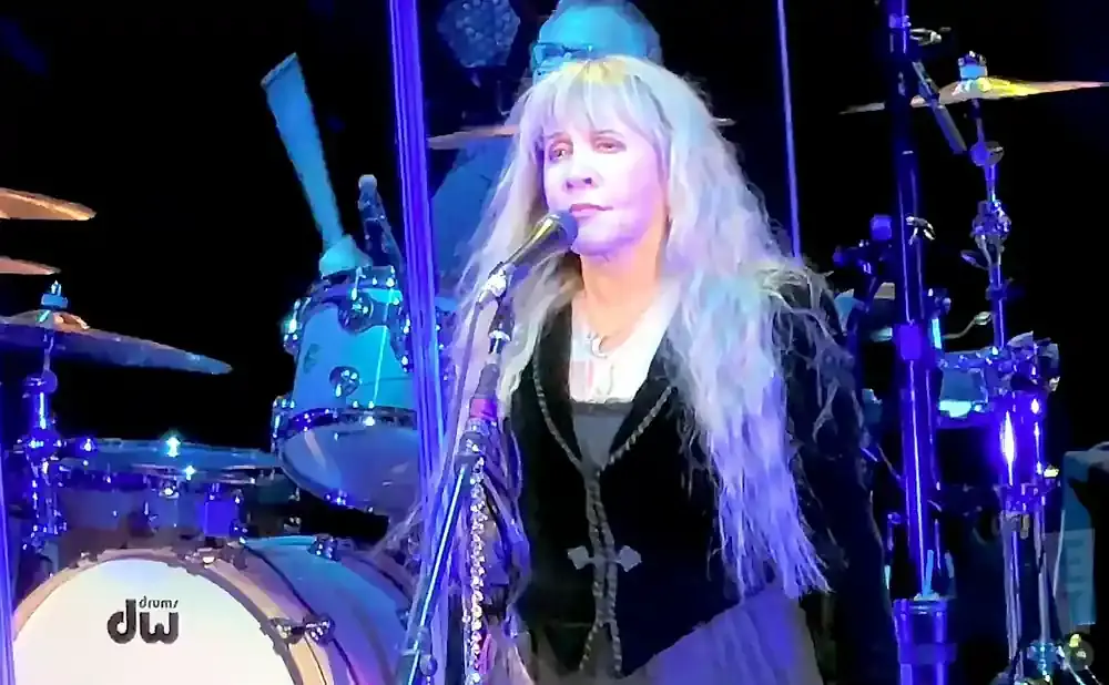 Stevie Nicks sings into a microphone on stage, with a drummer and drum kit visible behind her.