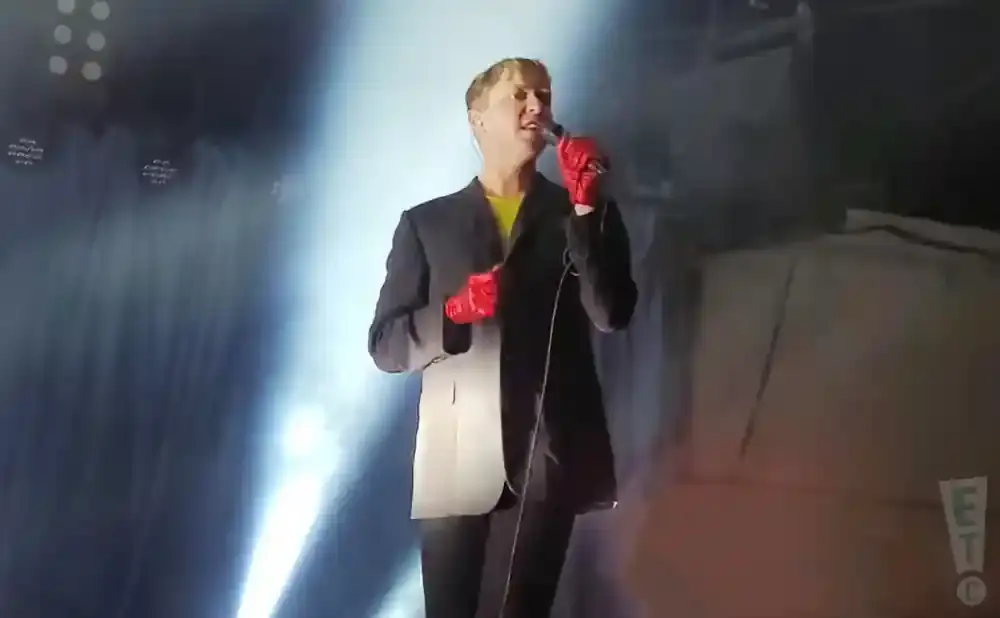 A person in a black suit and red gloves sings into a microphone on a stage with bright lights.