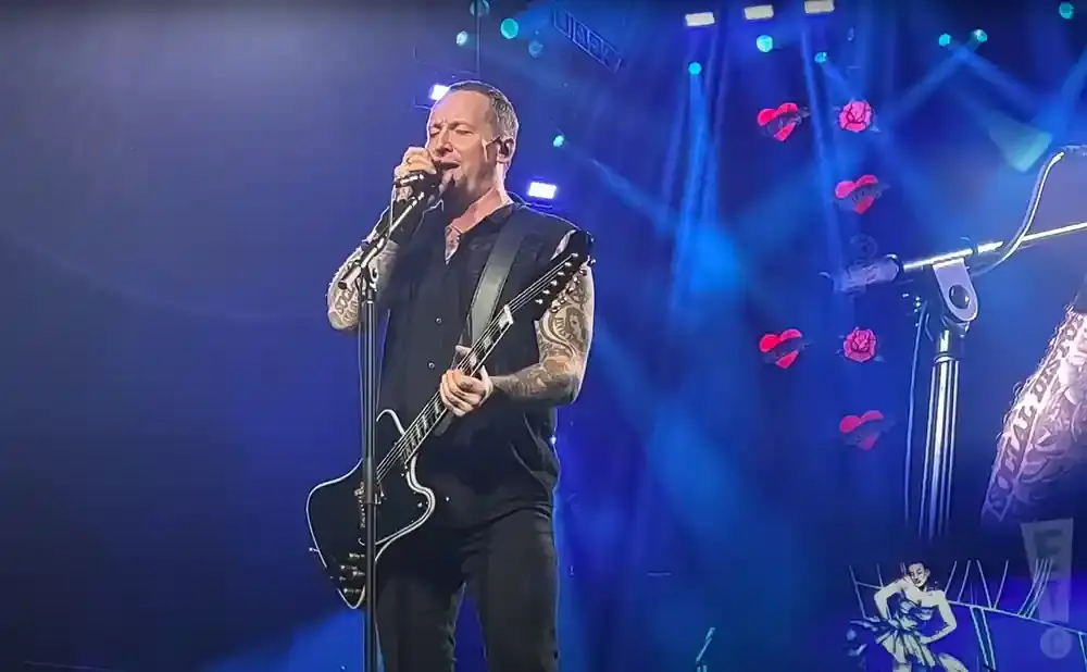 A man with tattoos sings into a microphone and plays a black electric guitar on a stage with blue lights and red heart decorations.