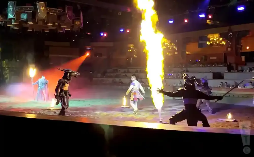 Performers in medieval costumes engage in a sword fight with flames on a stage.