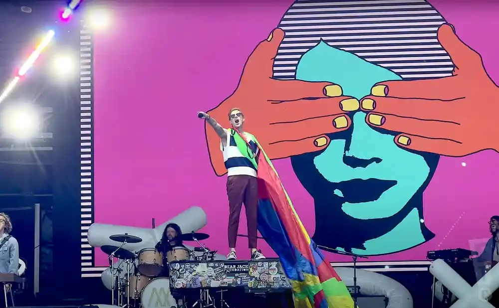 A performer in a striped shirt and colorful cape stands on a piano, pointing.