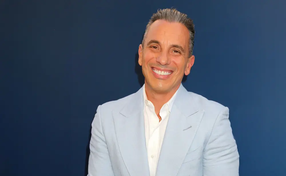 Comedian Sebastian Maniscalco smiles in a light blue suit and white shirt against a dark blue background.