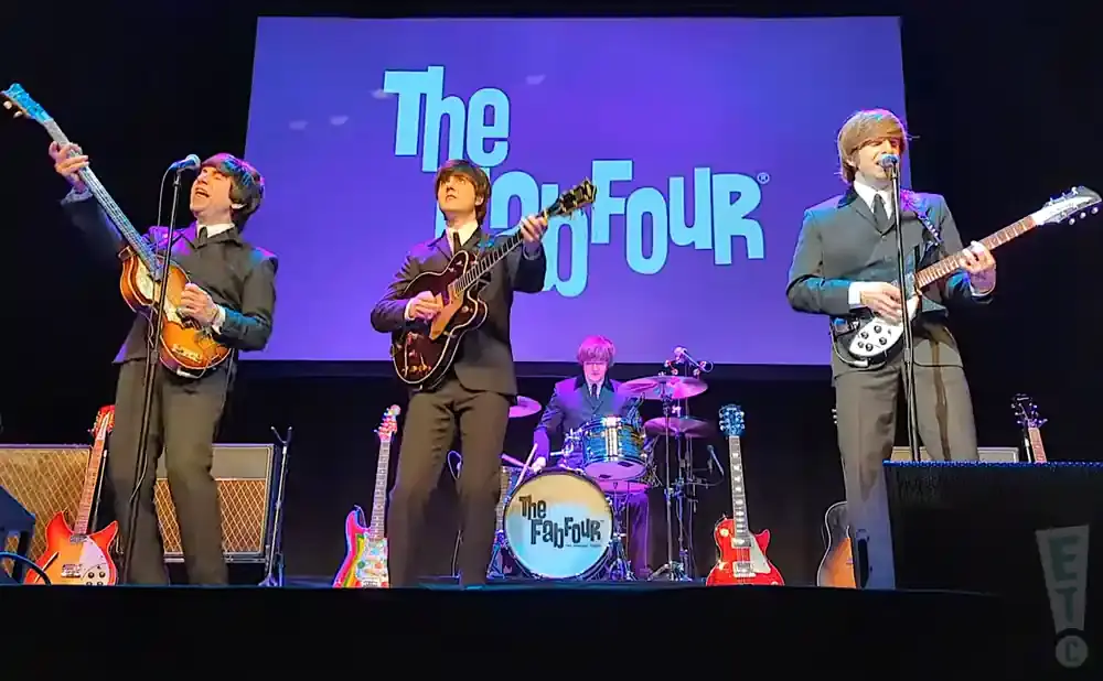 Four musicians dressed as The Beatles play guitars and drums on stage with 
