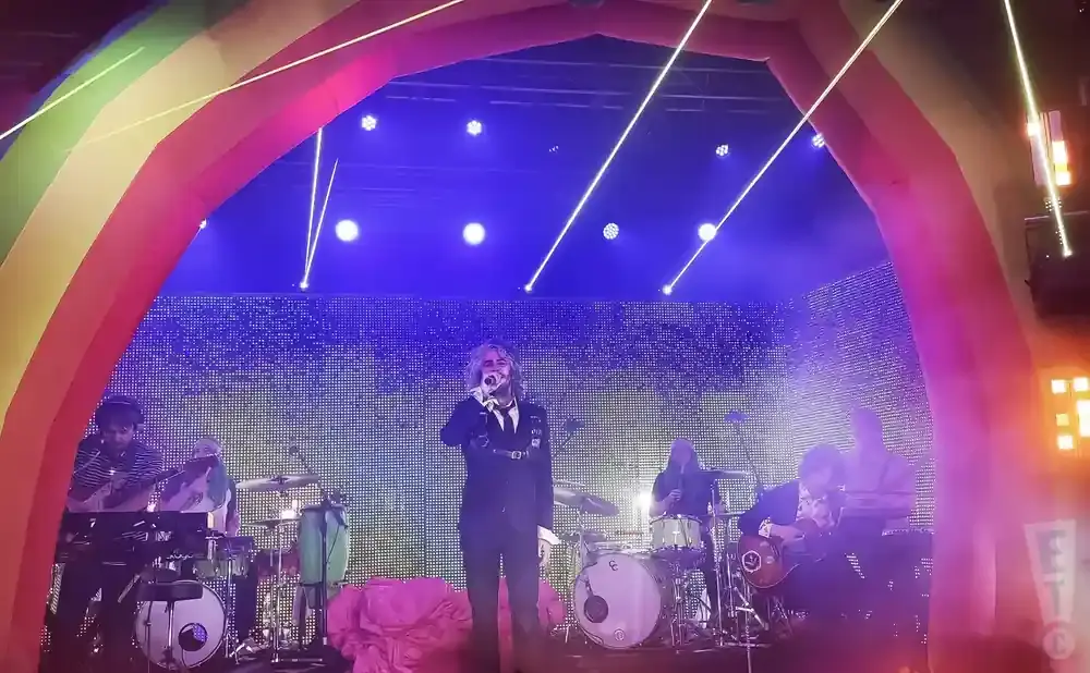 A band performs on a stage under a rainbow arch. The lead singer has long, wavy blonde hair and wears a black suit.