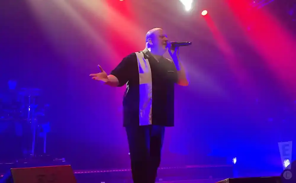 A man in a black shirt with a white stripe sings into a microphone on a stage with red and blue lights.