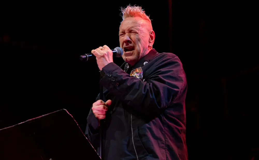 John Lydon sings into a microphone on stage, illuminated by red and blue lights.