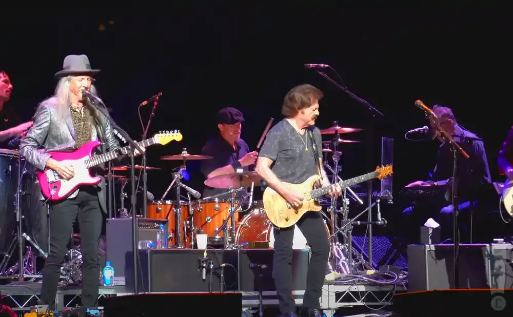 A band plays on a dimly lit stage. The guitarist in the foreground wears a sparkly jacket and hat.