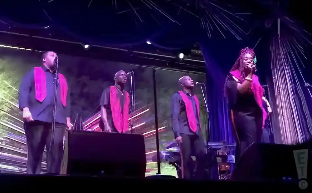 A group of singers on stage wearing pink robes over dark clothing.