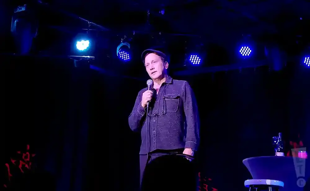 Comedian Bob Saget performs on stage, holding a microphone and wearing a dark shirt and hat.