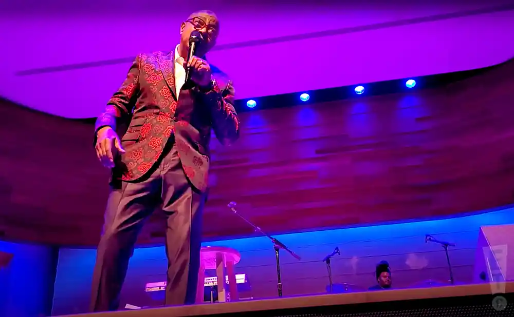 A Black man in a patterned suit sings into a microphone on a stage with purple lighting.