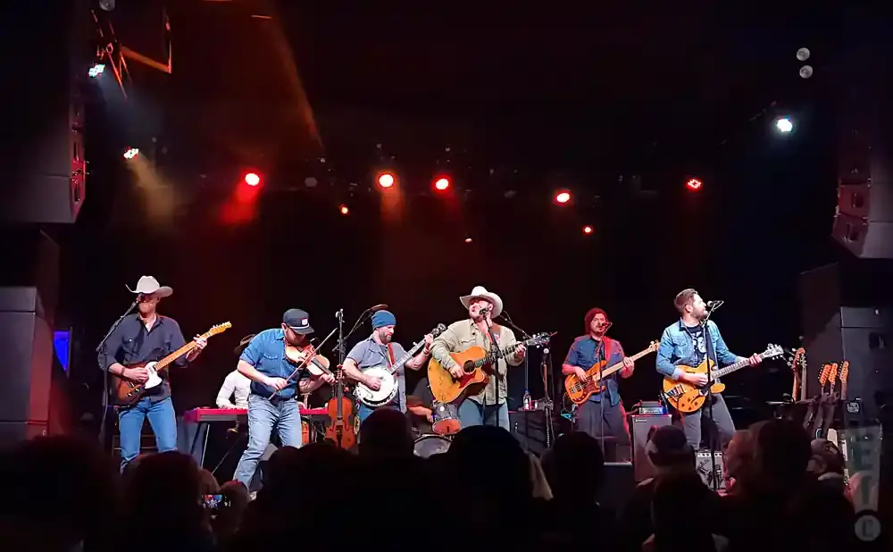 A band plays on a stage with red and white lights, instruments include guitars, banjo, and violin.