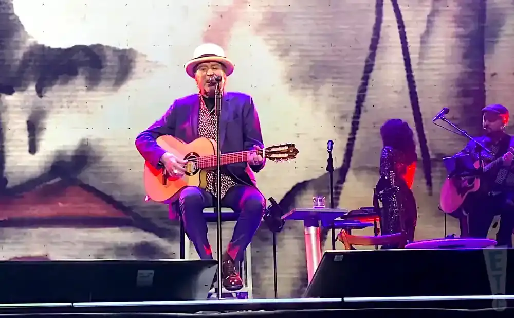 A man in a hat and suit plays guitar while singing on stage, with other musicians in the background.