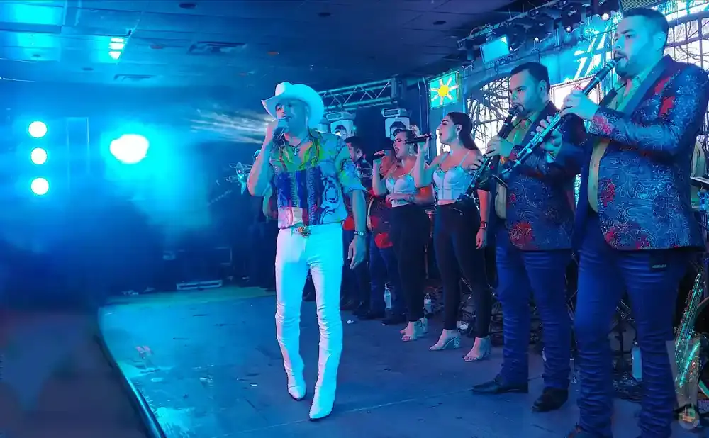 A band performs on stage with a male singer in a white cowboy hat and colorful shirt, two female singers, and two male clarinetists.