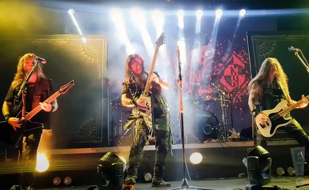 Three guitarists perform on stage with dramatic lighting.