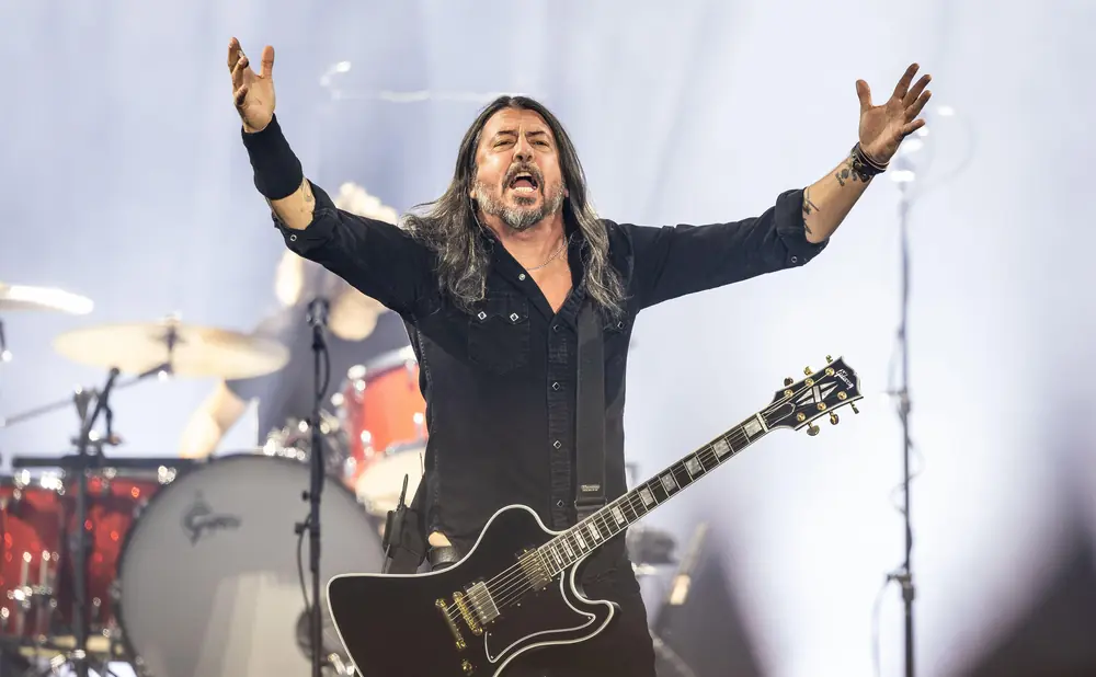 A man with long hair and a beard sings with his arms outstretched while playing a black electric guitar.