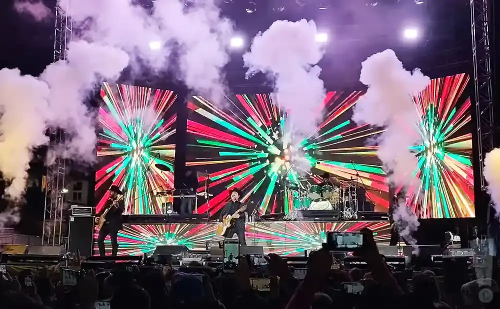Band performing on a stage with a colorful, abstract light show backdrop and smoke effects.