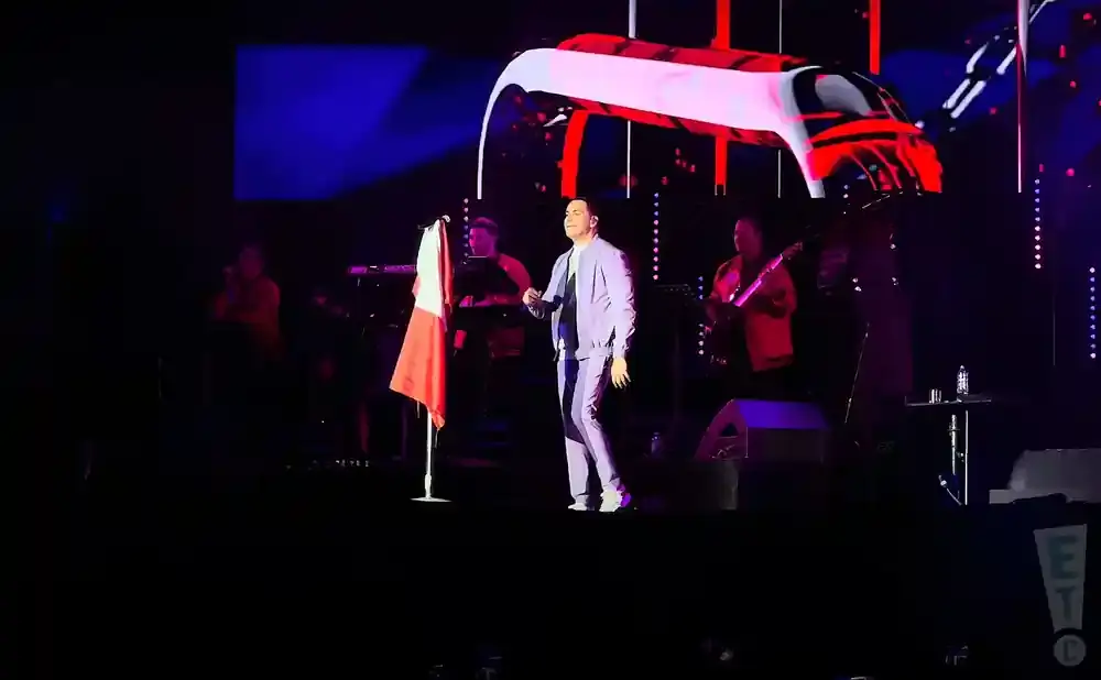 A performer in a light blue suit stands on stage with a band, holding a flag.
