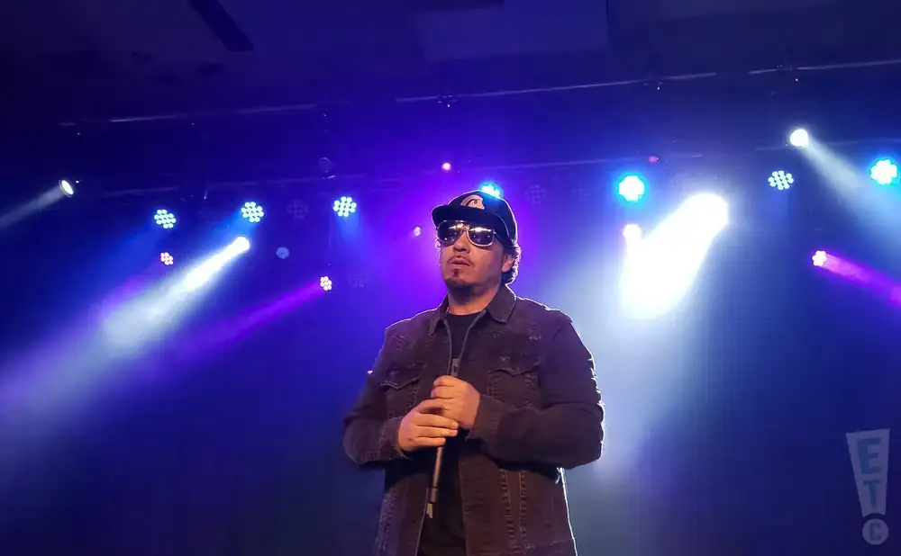 A man in sunglasses and a baseball cap performs on a stage with purple and white lights.